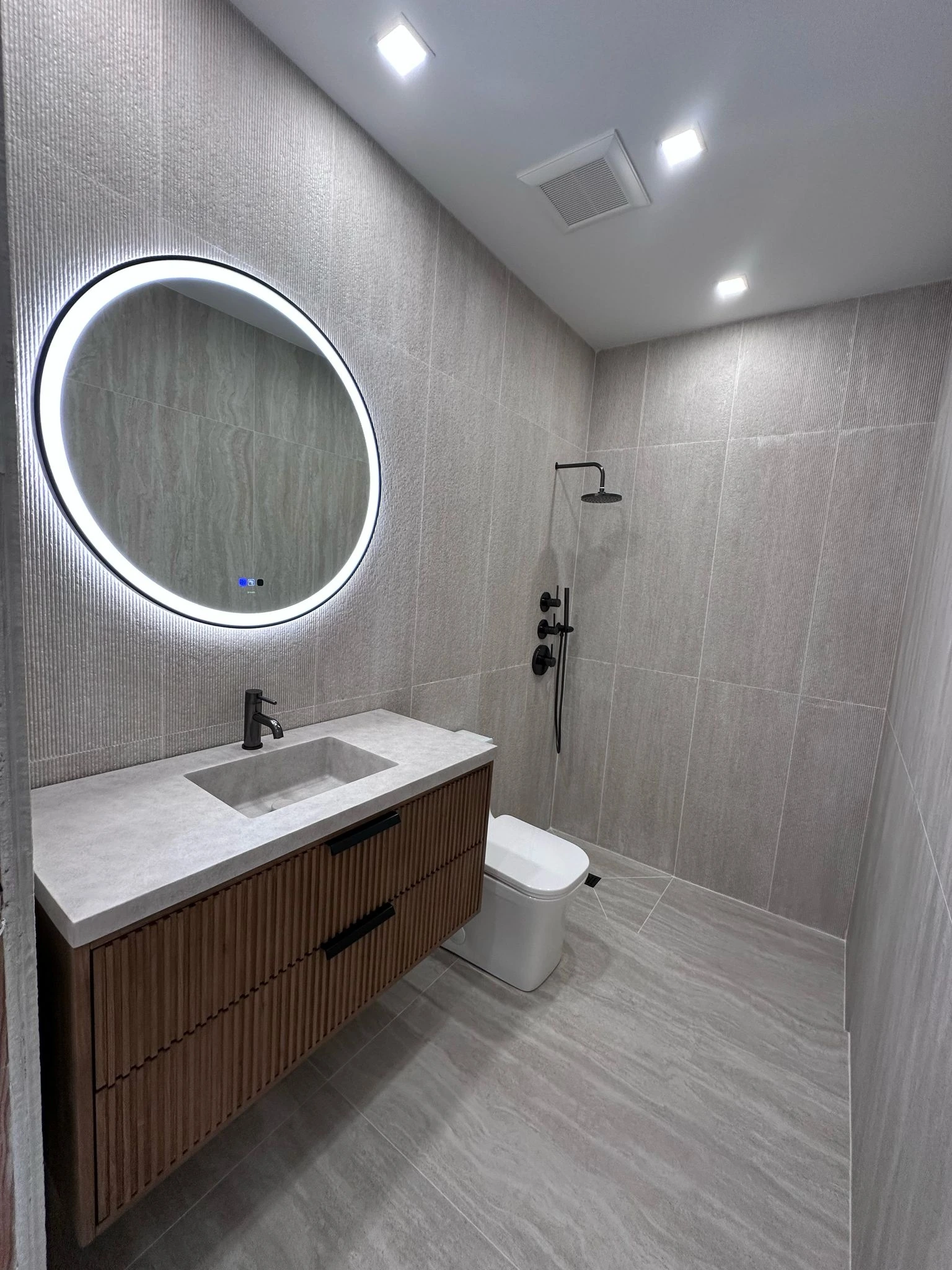 Grey tile bathroom with round backlit mirror and floating wood vanity — Plantation, FL